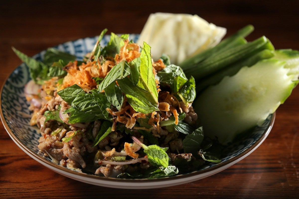 A plate of Thai larb salad featuring minced meat mixed with herbs, sliced shallots, and chili flakes, topped with fresh mint leaves and crispy fried shallots. The dish is accompanied by fresh cucumber slices, long green beans, and cabbage wedges on the side. The plate has a blue and white pattern and is set on a wooden surface.