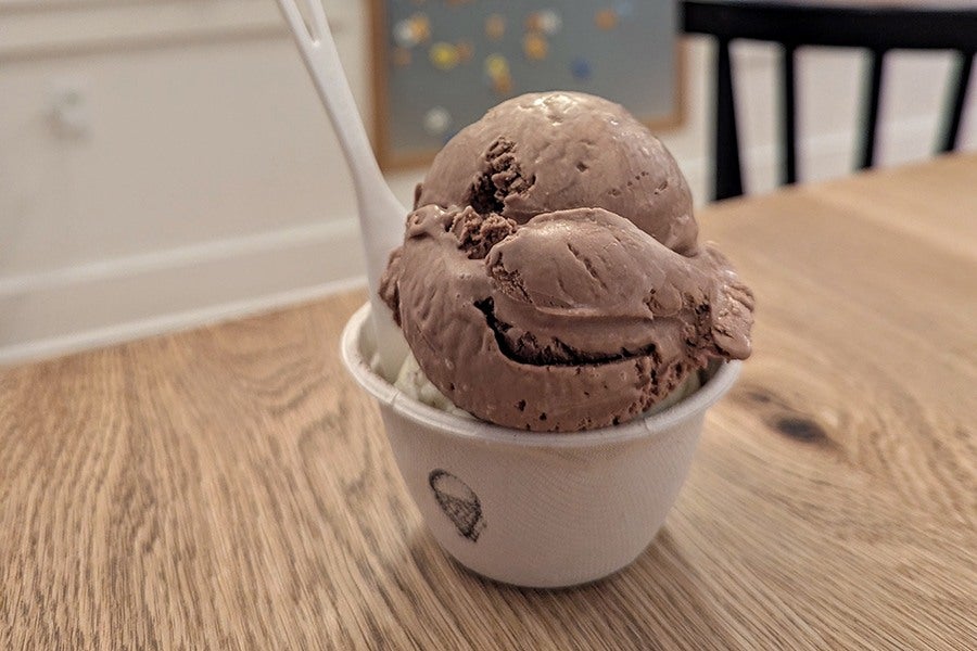 Two scoops of chocolate ice cream in a small white paper cup with a white plastic spoon, placed on a wooden table.