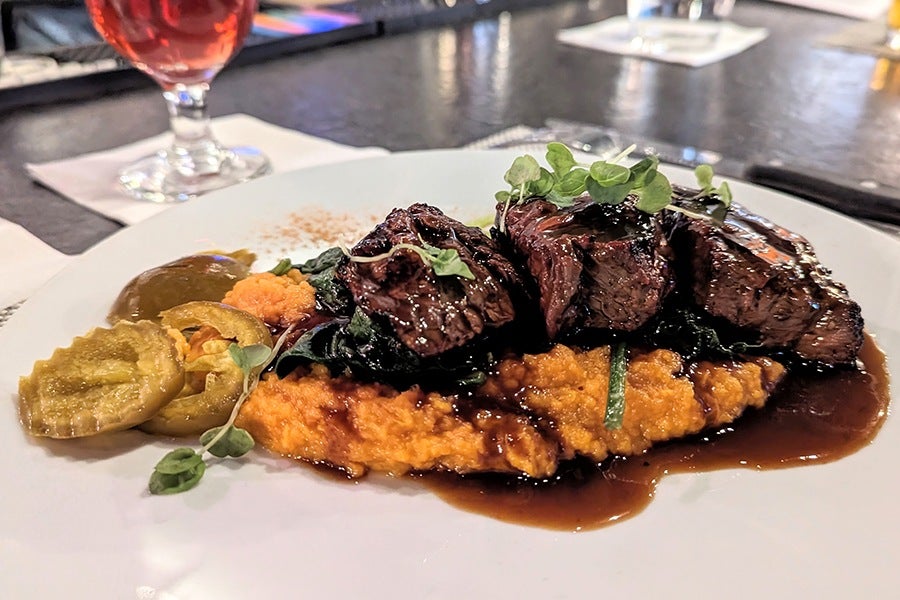Grilled steak slices served on a bed of creamy orange mashed sweet potatoes, topped with sautéed greens and microgreens, accompanied by pickled jalapeño slices and drizzled with a rich brown sauce.