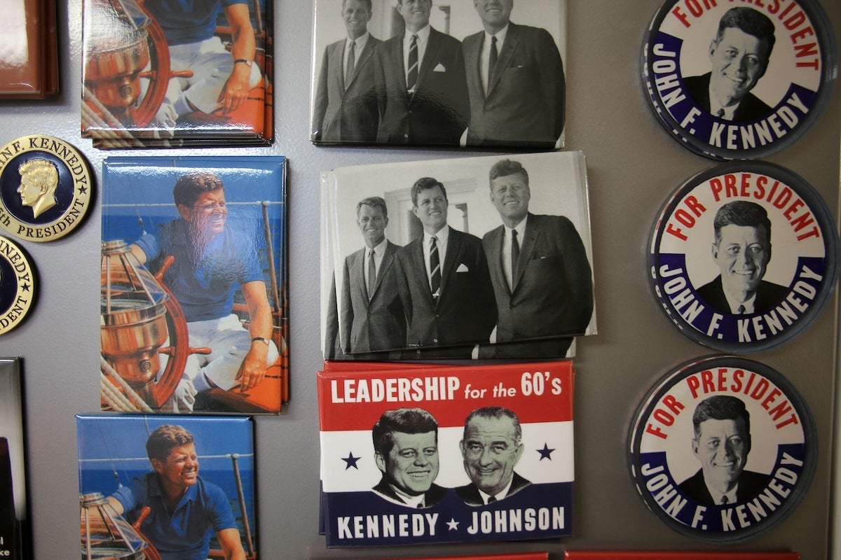 A collection of vintage political memorabilia featuring John F. Kennedy. The items include campaign buttons with "For President John F. Kennedy" text and his portrait, rectangular pins with images of Kennedy in casual attire at a ship's wheel, black-and-white photos of Kennedy with other men in suits, and a rectangular pin with "Leadership for the 60's Kennedy Johnson" text and portraits of John F. Kennedy and Lyndon B. Johnson.