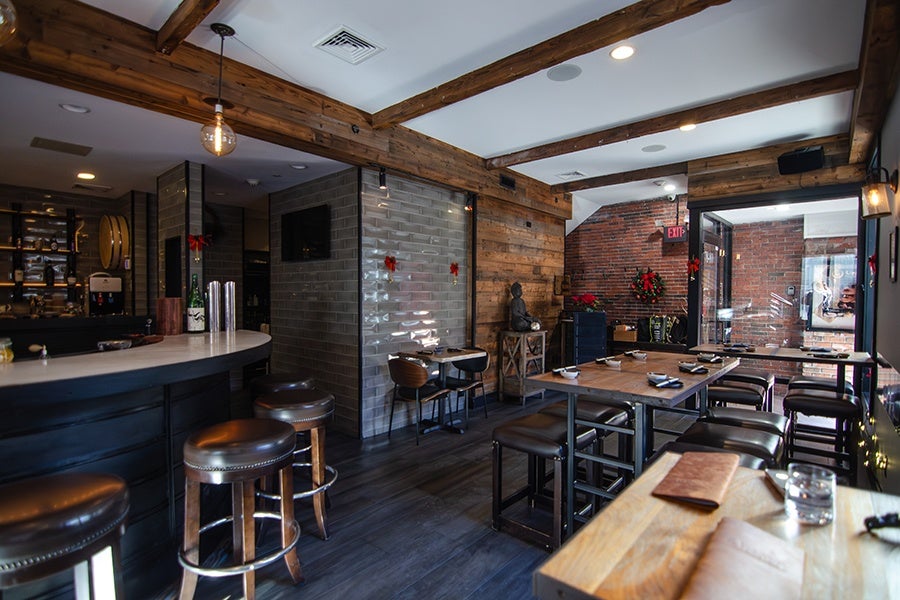 A cozy restaurant interior featuring wooden beams on the ceiling and a mix of brick and wood-paneled walls. There is a curved bar with leather-topped stools on the left and several wooden tables with matching stools arranged on the right. The space is softly lit with hanging light bulbs and recessed ceiling lights. Decorative elements include a small statue on a side table, a wreath on the brick wall, and red bows on the walls. The floor is dark wood, complementing the warm, rustic atmosphere.
