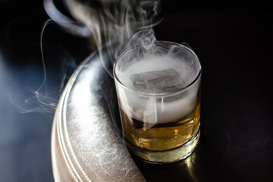A glass of amber-colored bourbon with a large ice cube, emitting swirling white smoke, placed on a dark surface next to a leather edge with visible stitching.