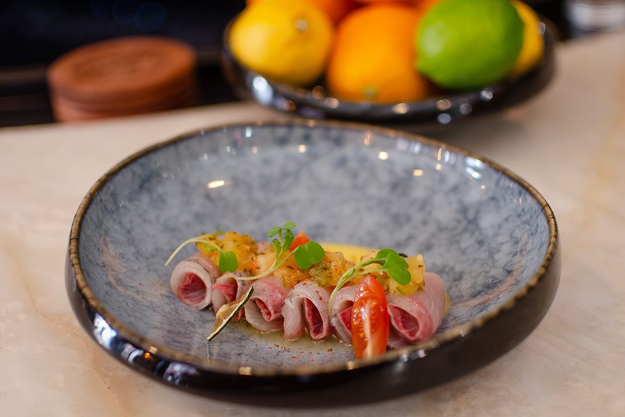 A dish featuring rolled slices of raw fish, garnished with small green microgreens, a slice of cherry tomato, and a topping of finely chopped yellow rsalsa, all presented on a blue-gray ceramic plate with a dark rim. In the background, there is a blurred bowl of assorted citrus fruits including lemons, oranges, and a lime.