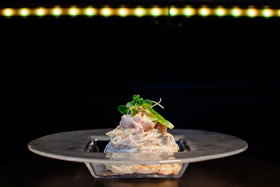 A serving of white noodles garnished with small green leaves and a slice of green vegetable, presented on a clear glass plate with a dark background and a row of yellow lights above.