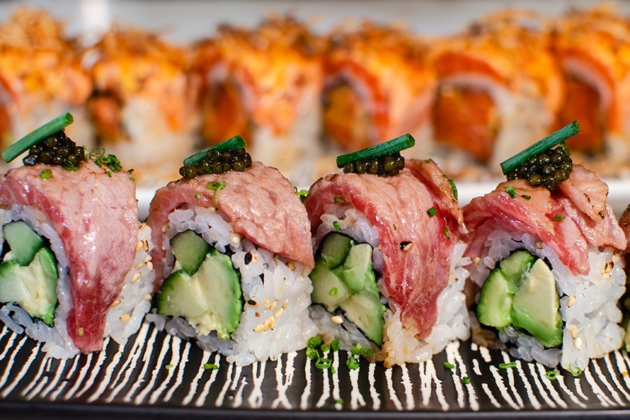 Four pieces of sushi rolls topped with thin slices of seared tuna, small dollops of black caviar, and green garnish. The rolls contain cucumber and avocado, wrapped in white rice with sesame seeds. In the background, there is a blurred view of additional sushi rolls with an orange topping. The sushi is presented on a black and white striped plate.