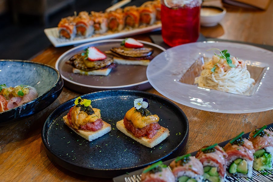 Two pieces of sushi topped with sea urchin, caviar, and edible flowers on small rectangular rice bases, served on a dark blue plate. Surrounding dishes include sushi rolls, a plate with two pieces of food topped with sliced strawberries, a bowl of noodles garnished with herbs, and a blue bowl with sashimi. A red drink in a glass jar is also visible.