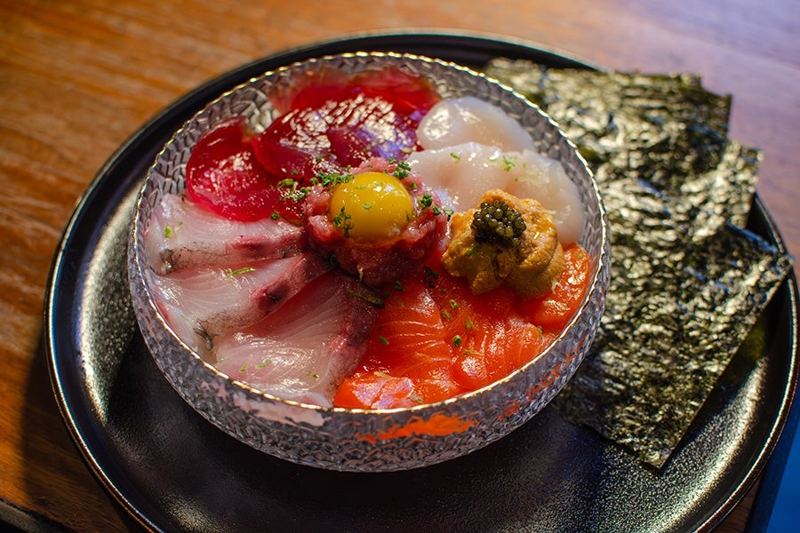 Slices of various raw fish are arranged in a bowl, topped with a raw quail egg and a dollop of caviar-topped sea urchin. The bowl sits on a black plate with a stack of seaweed sheets alongside.
