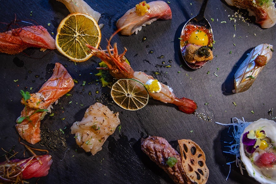 An assortment of sushi and sashimi pieces artfully arranged on a dark slate surface. The selection includes various types of fish, shrimp, and garnishes such as dried citrus slices, herbs, and edible flowers. One piece is served on a spoon with vibrant toppings, and another features a lotus root chip. The presentation is colorful and elegant, highlighting the freshness and variety of the seafood.