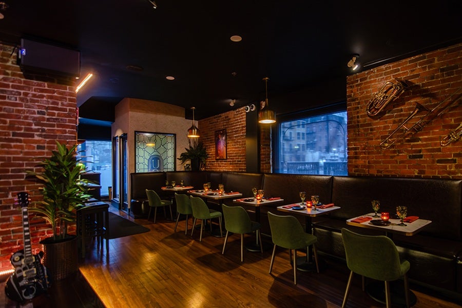 Cozy restaurant interior with exposed brick walls, dark ceiling, and warm lighting. There are several tables with green upholstered chairs and black leather bench seating along the wall. Each table is set with glasses, napkins, and small candles. Brass musical instruments are mounted on the brick wall as decoration, and a guitar is placed near a potted plant on the left side. Large windows show a blurred cityscape outside.