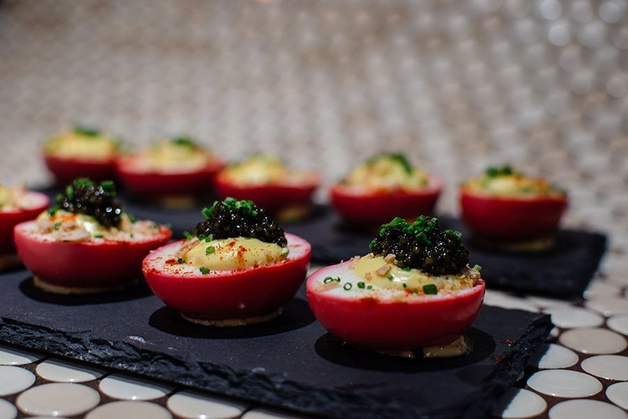 Red-tinted deviled eggs filled with a creamy yellow mixture, topped with black caviar and garnished with chopped green herbs, arranged on black slate serving trays. The background features a blurred pattern of circular tiles.