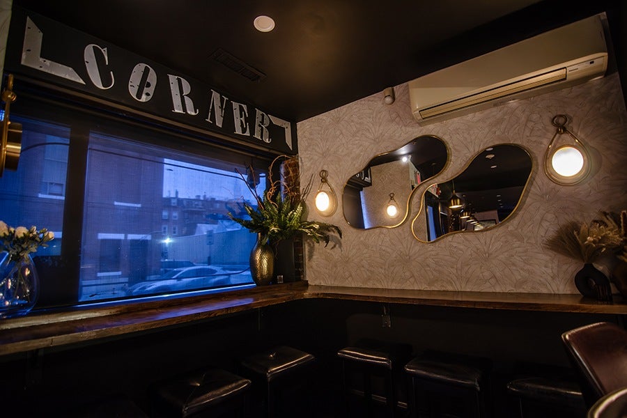 Cozy corner seating area with a wooden countertop along the walls, three black cushioned stools, two abstract-shaped mirrors with gold frames, two round wall lights with gold accents, a large window with a blue tint, and decorative vases with plants and flowers. An air conditioning unit is mounted on the wall above the mirrors. The word "CORNER" is displayed in large, white, vintage-style letters on a black background above the window.