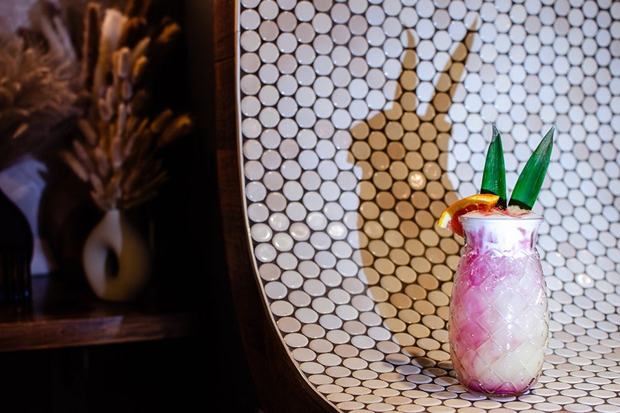 A pink and white cocktail garnished with a citrus slice is served in a glass that looks like a pineapple, garnished with two tall green leaves. It projects a rabbit-like shadow on the white tiled wall behind it.
