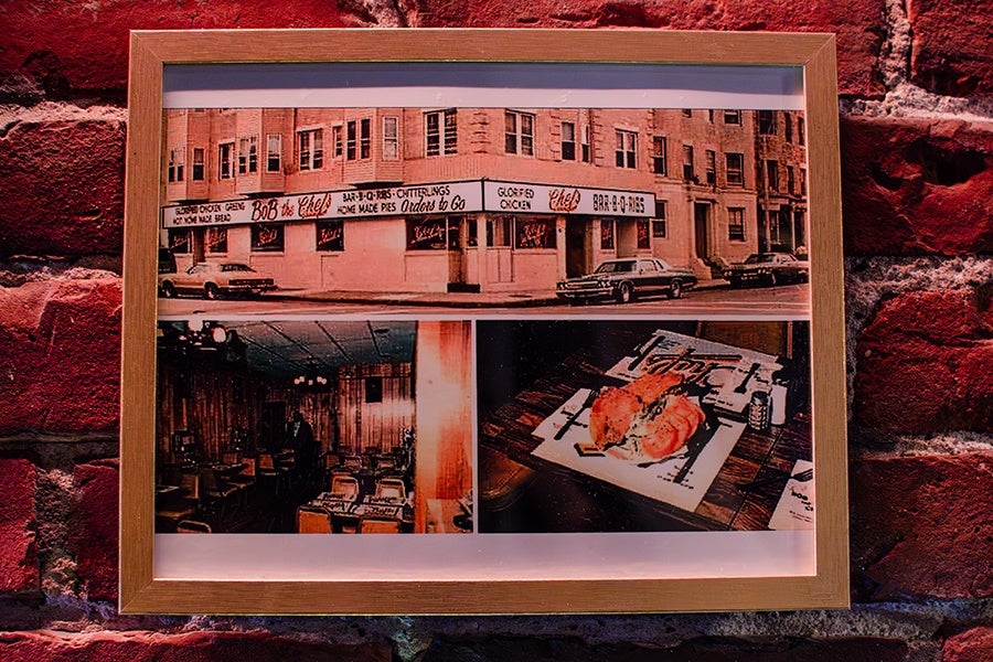 Framed collage featuring three vintage photos: the top photo shows the exterior of a restaurant named "Bob the Chef" with classic cars parked outside; the bottom left photo depicts the restaurant's interior with wooden paneling and tables set for dining; the bottom right photo displays a close-up of a sandwich on a table with utensils and a drink. The frame is wooden and mounted on a red brick wall.