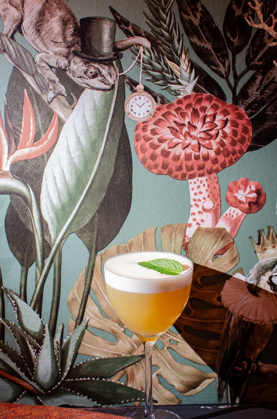 A yellow cocktail with a frothy white top, garnished with a fresh mint leaf, served in a stemmed glass. The background features a colorful mural with large leaves, red mushrooms, and a chameleon wearing a top hat holding a pocket watch.