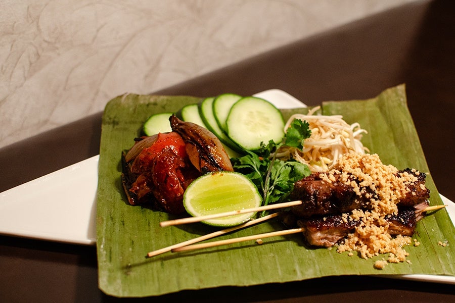 Two grilled meat skewers topped with crushed peanuts, served on a banana leaf with sliced cucumber, lime half, fresh herbs, bean sprouts, and roasted vegetables.
