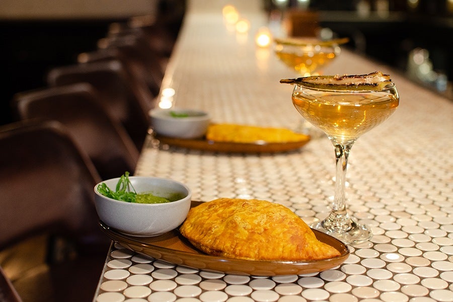 A brown plate with a golden-brown beef or chicken patty and a small bowl of green sauce garnished with herbs, placed on a white tiled bar counter. Next to the plate is a coupe glass filled with a light amber cocktail, garnished with charred, pickled okra. Additional plates and glasses are visible further down the counter.