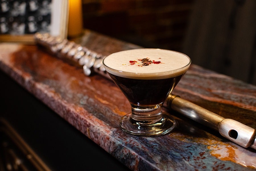 A glass of dark espresso martini with a creamy foam top garnished with small red and brown flakes, placed on a polished stone countertop next to a flute.