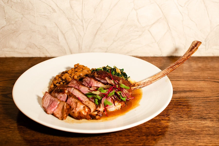 Sliced cooked veal chop garnished with microgreens, served with a side of cooked beans and sautéed greens on a white plate, placed on a wooden table.