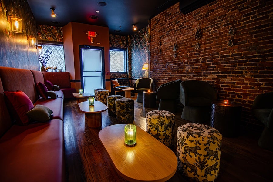 Cozy lounge area with a long maroon sofa on the left, several patterned ottomans, dark armchairs, and wooden coffee tables. The room features exposed brick walls, dim warm lighting from wall sconces and a table lamp, and decorative elements including hanging wall pieces and floral wallpaper near the entrance door.