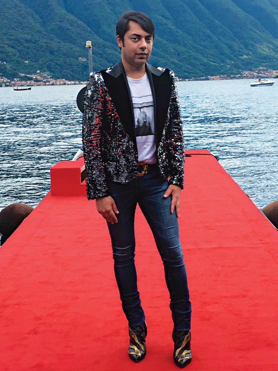 A person stands on a red carpeted dock by a body of water with mountains in the background. They are wearing a black and silver sequined blazer over a white graphic t-shirt, dark skinny jeans, and black and gold pointed-toe boots.