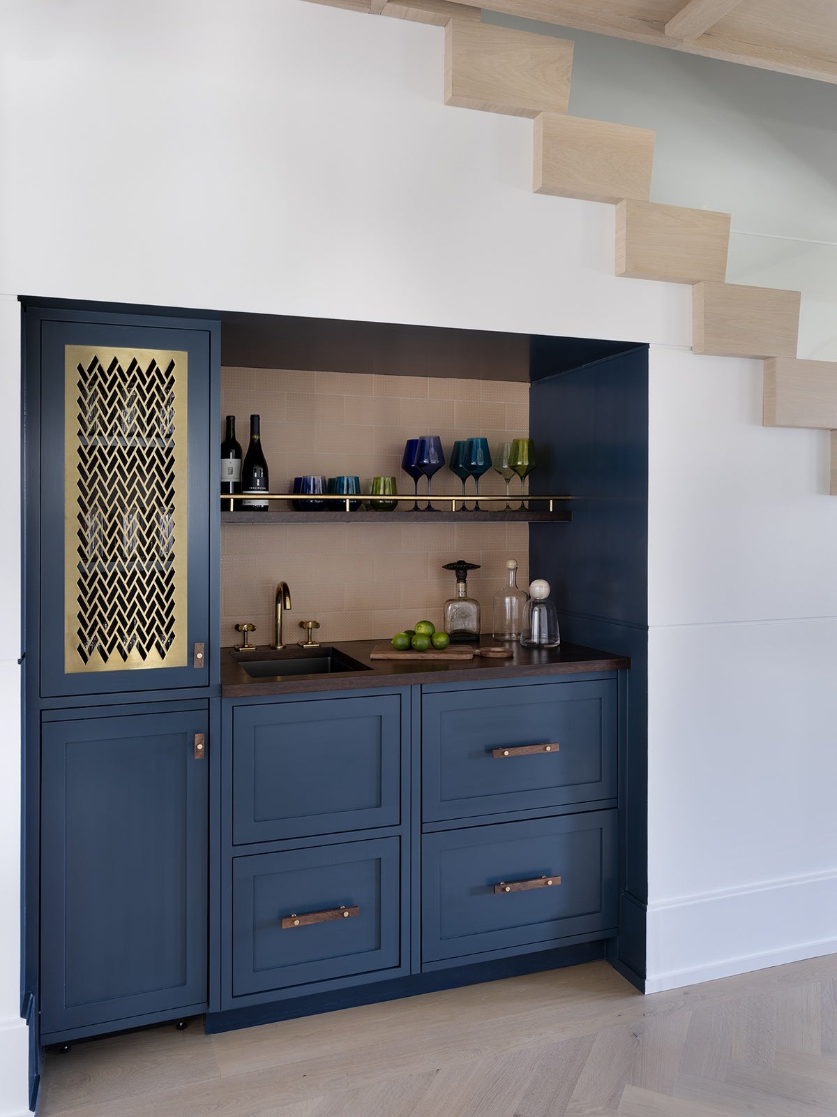 A built-in wet bar with navy blue cabinetry and a dark wood countertop. The left cabinet door features a decorative gold metal grille. The bar includes a small sink with brass fixtures, a shelf holding colorful glassware, and a few bottles. The backsplash is a light beige tile. The wet bar is set under a staircase with light wood steps and white walls.