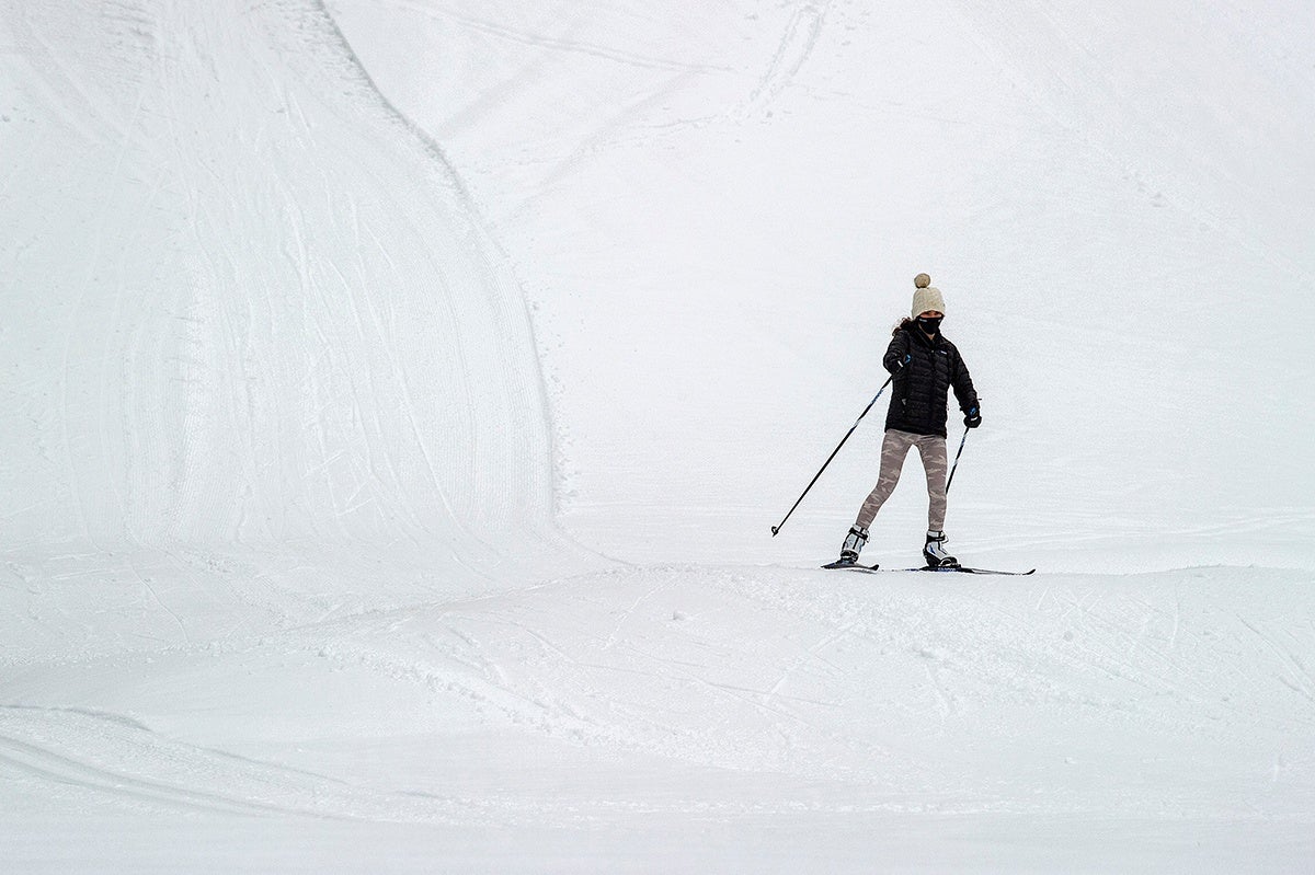 A person skiing on a snowy slope wearing a black jacket, beige patterned pants, black gloves, a beige knit hat with a pom-pom, and a black face mask, holding ski poles.