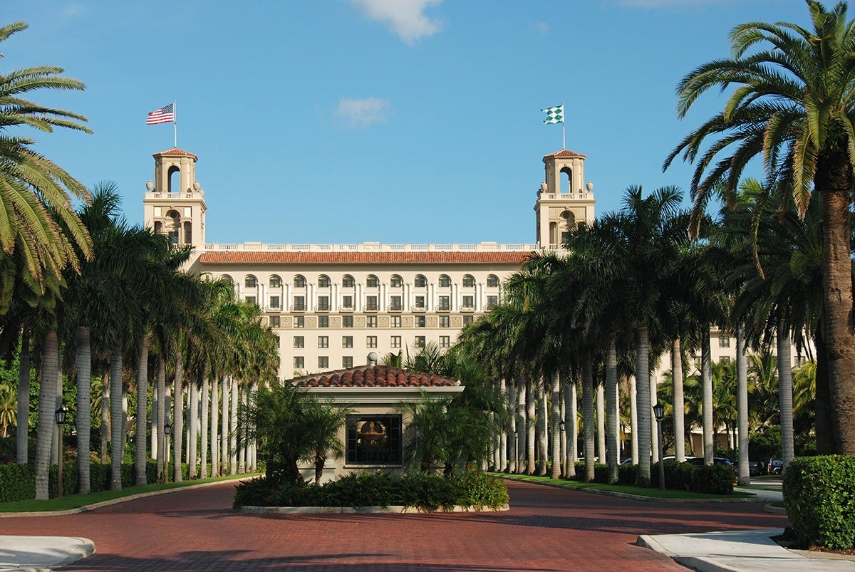 A large, elegant building with two towers, each topped with a flag, is framed by symmetrical rows of tall palm trees on either side of a red brick driveway. The building has multiple windows and a tiled roof, with a small guardhouse or entrance structure in the center of the driveway surrounded by greenery. The sky is clear and blue.