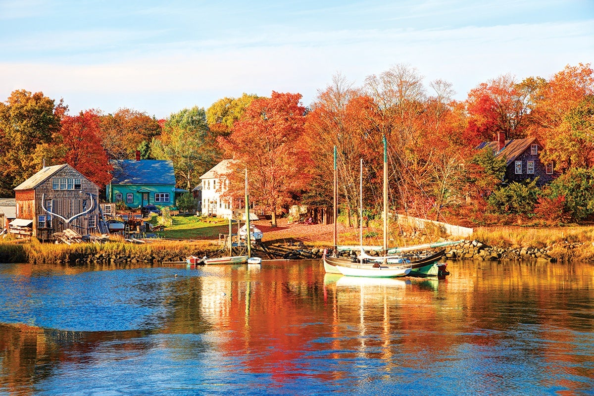 A calm waterfront scene in autumn with colorful fall foliage in shades of orange, red, and yellow. Several small houses and buildings, including a rustic wooden boathouse with large antlers mounted on the front, line the shore. Two sailboats are moored in the water, reflecting the vibrant colors of the trees and sky. The sky is clear with soft clouds, and the overall atmosphere is peaceful and picturesque.