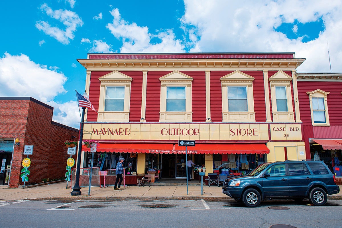 A two-story red and cream building with a sign reading "Maynard Outdoor Store" and "Case Bld. 28" on the front. The building has three large windows on the second floor with decorative trim. Below, a red awning extends over the sidewalk, shading the store entrance and display area. A blue SUV is parked on the street in front, and two people are walking on the sidewalk. An American flag is mounted on a lamppost near the left side of the building. The sky is bright blue with scattered white clouds.
