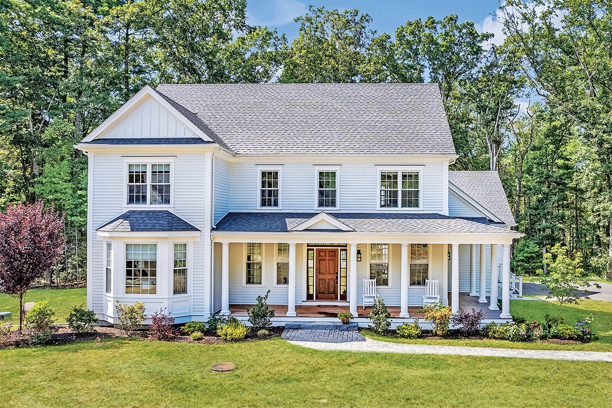 Two-story white house with a gray shingled roof, featuring a covered front porch supported by white columns. The porch has two white rocking chairs and a wooden front door with sidelights. The house has multiple windows, including a bay window on the left side. The front yard is landscaped with shrubs, small trees, and a curved stone pathway leading to the porch steps. Tall trees surround the property in the background.