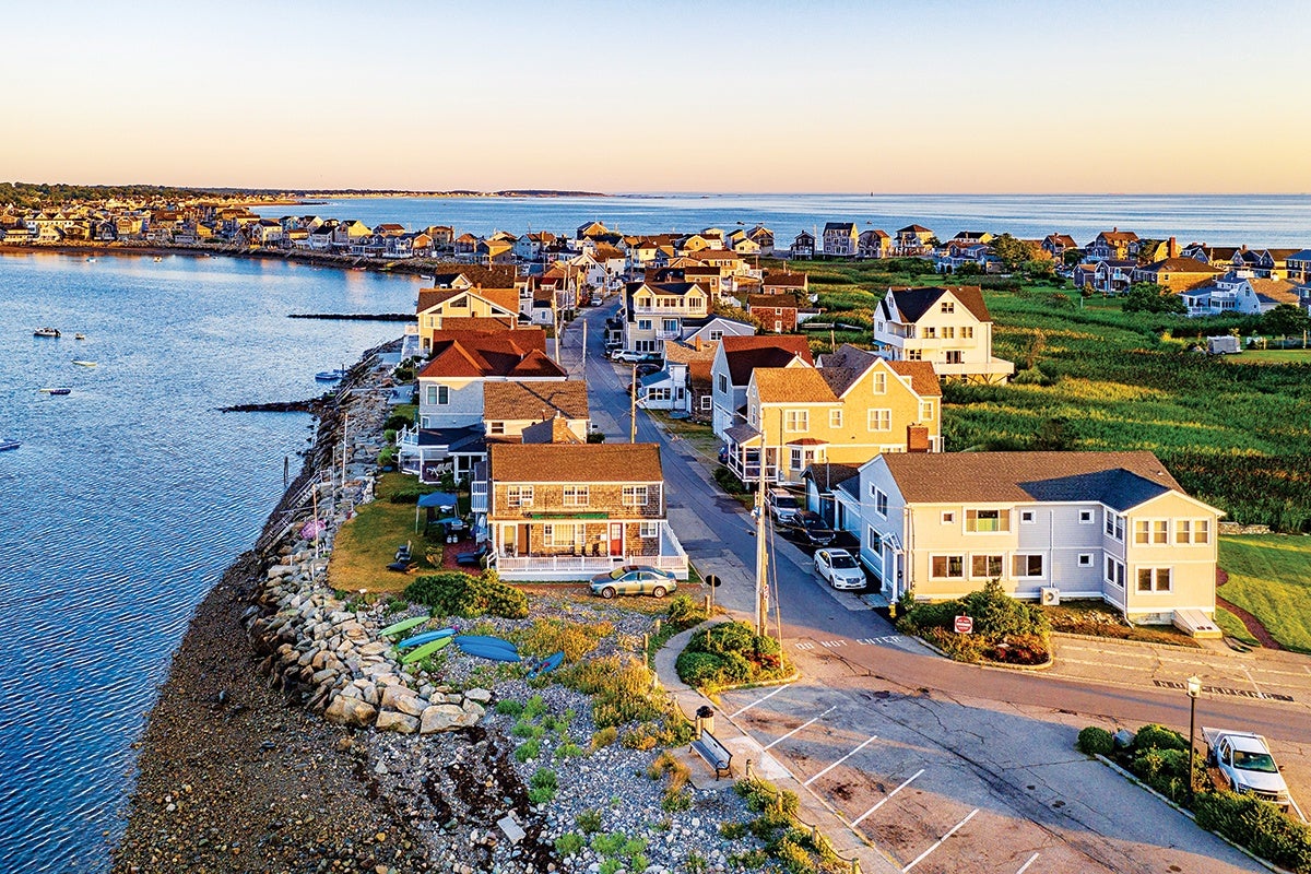 A coastal neighborhood with rows of houses along a narrow strip of land bordered by water on the left and green fields on the right. The houses are mostly two-story with varied roof styles and colors. A paved road runs through the middle, with parked cars visible. The shoreline features rocky edges and several kayaks or small boats resting on the ground near the water. The scene is bathed in warm, golden sunlight, suggesting early morning or late afternoon.