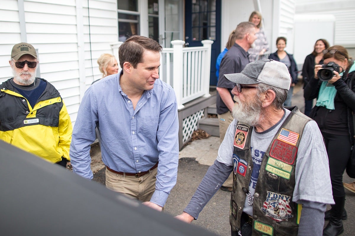 A man in a light blue button-up shirt and beige pants is shaking hands with an older man wearing a gray cap, glasses, a gray long-sleeve shirt, and a black vest adorned with various patches, including U.S. Army and FDNY insignias. Another man in a yellow and black jacket with a "Veteran" cap stands nearby. Several people, including a woman with a camera, are in the background outside a white building with a porch.