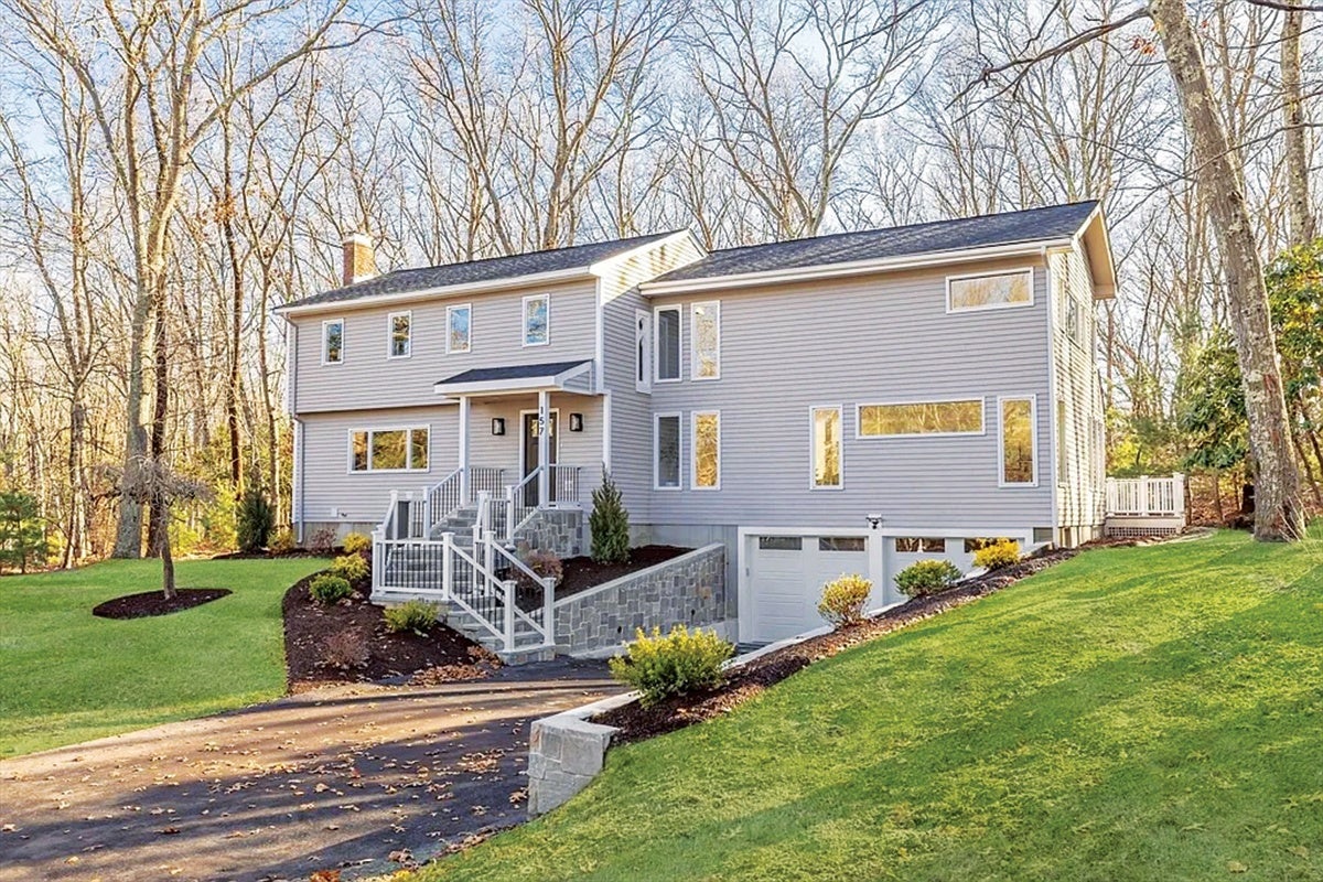 Two-story modern house with light gray siding and multiple rectangular windows, surrounded by a green lawn and leafless trees in the background. The house features a stone staircase with white railings leading to the front door and a driveway extending to a garage on the right side.