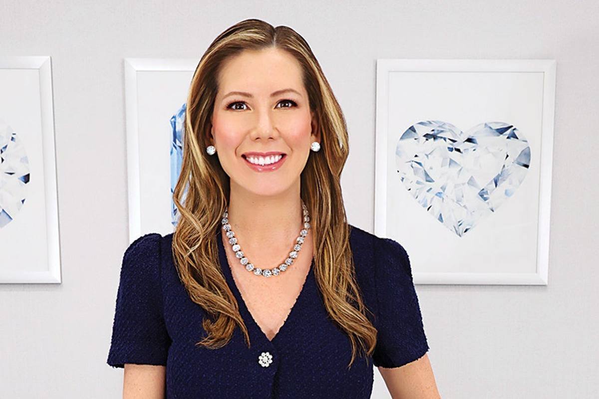 A woman with long, light brown hair wearing a navy blue textured dress with a decorative button. She has a diamond necklace and matching diamond stud earrings. Behind her are two framed pictures of large, faceted diamonds, one shaped like a heart. The background wall is light gray.