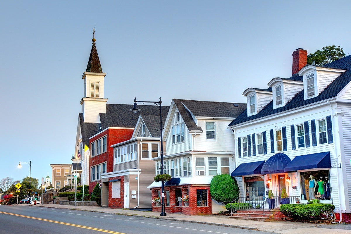 A street view of a small town with a row of buildings including a white church with a tall steeple, a red brick building, and several white houses with blue shutters and awnings. The sky is clear and the street is empty.
