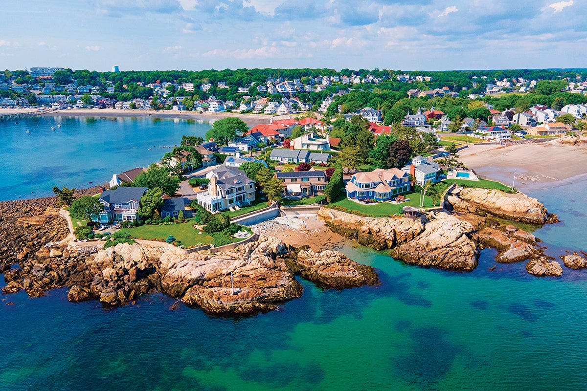 A coastal residential area with large houses situated on rocky cliffs overlooking clear blue-green ocean water. The homes have well-maintained lawns and gardens, and the background shows a densely built neighborhood with numerous houses and trees under a partly cloudy sky.