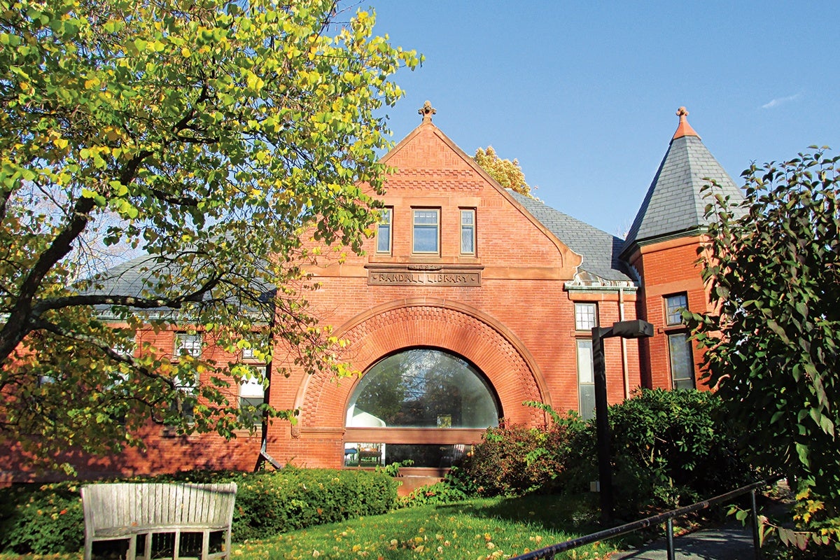 Red brick building with a large arched window in the center, labeled "Randall Library." The building has a steep gray slate roof and a turret on the right side. Green bushes and trees with yellowing leaves surround the building, and a wooden bench is visible on the left side. The sky is clear and blue.