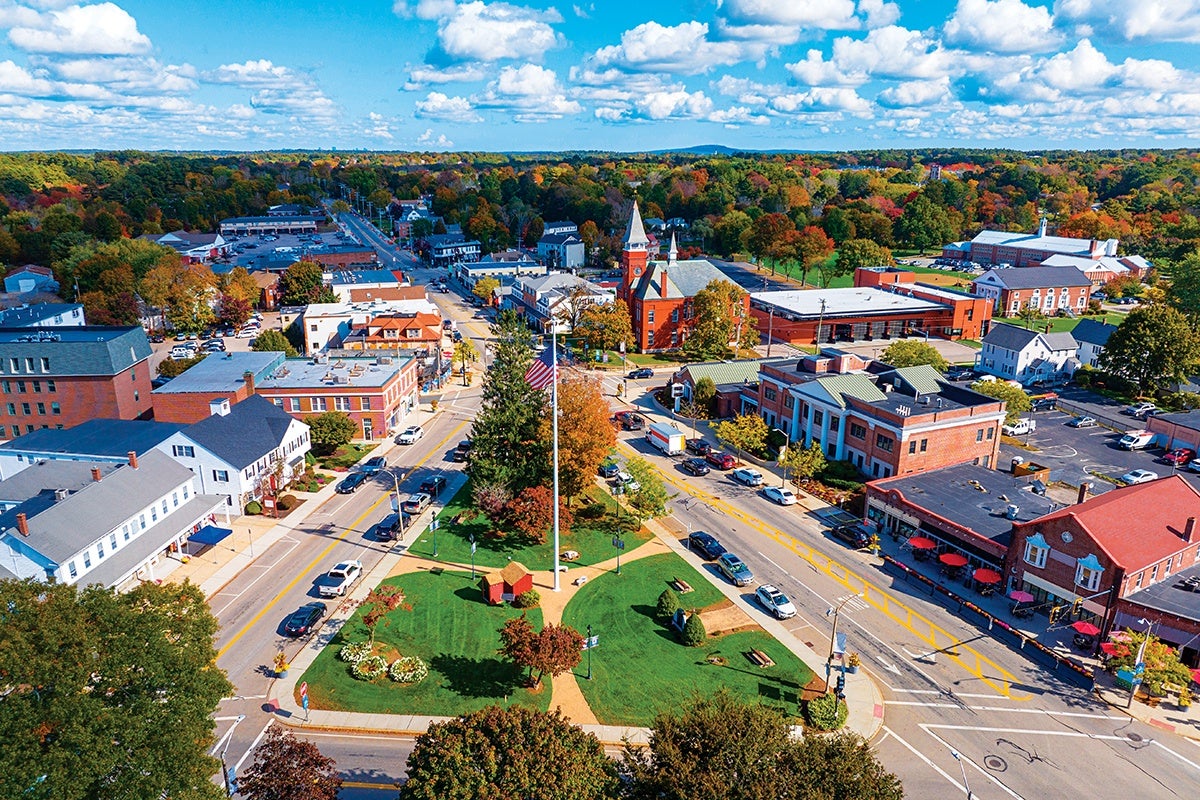A small town center with a triangular green park in the middle featuring a tall flagpole with an American flag. Surrounding the park are streets with parked cars and buildings including brick structures, a church with a steeple, and various commercial and residential buildings. Trees with autumn foliage and a partly cloudy blue sky are visible in the background.