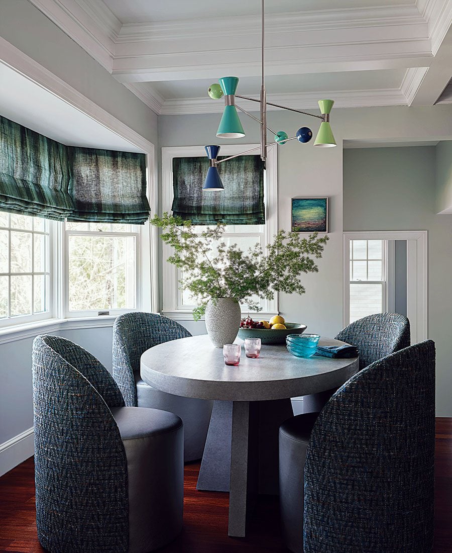 Round dining table with a gray stone-like surface surrounded by four upholstered chairs in a textured blue fabric. A large vase with green leafy branches sits in the center of the table, accompanied by a bowl of fruit, two small pink glasses, and a blue glass bowl with a folded napkin. The room features large windows with dark green Roman shades, white trim, and a modern ceiling light fixture with blue and green cone-shaped shades. The floor is dark wood, and the walls are painted light gray.