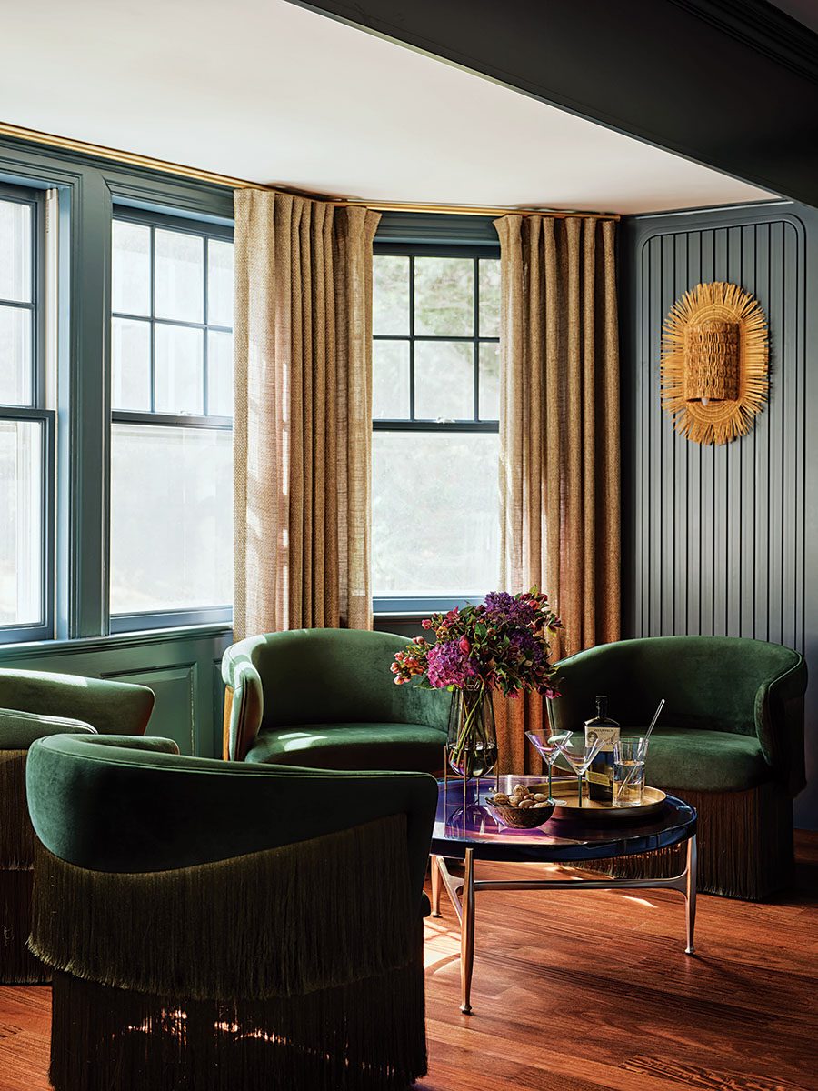 A cozy corner seating area features three dark green velvet armchairs with fringe detailing along the bottom. They surround a round glass coffee table with a gold tray holding a bottle, two martini glasses, and a tumbler with a straw. A vase with purple and pink flowers adds a vibrant touch. The room has large windows framed with blue trim and beige curtains, and the walls are painted a muted blue with vertical paneling. A decorative woven wall sconce in natural tones is mounted on the wall. The floor is a warm-toned wood.