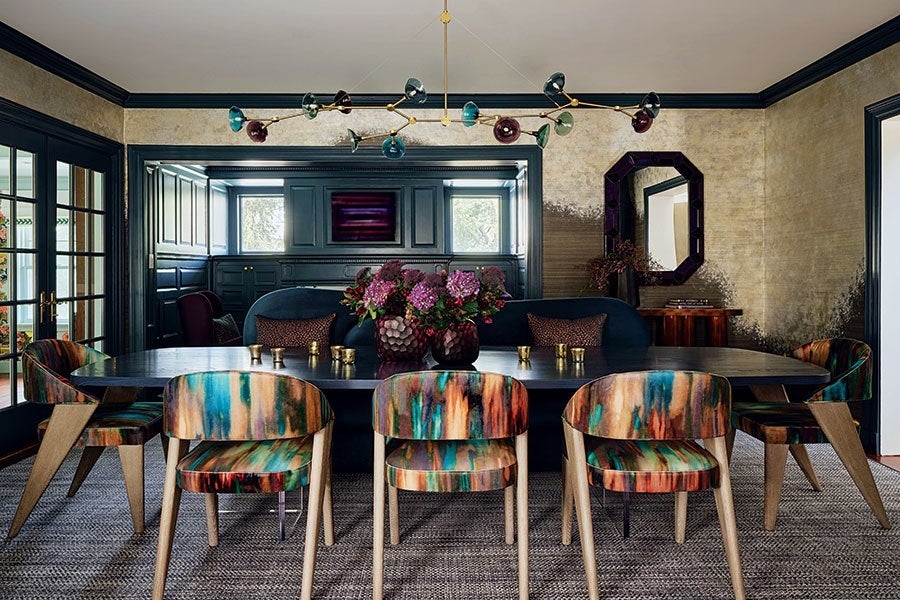 Dining room with a large dark rectangular table surrounded by eight colorful, abstract-patterned chairs with wooden legs. A dark blue upholstered bench with patterned pillows is positioned against the back wall. Two vases with purple flowers are centered on the table. The room features dark wood paneling, a textured beige and black wall, a geometric mirror, and a modern chandelier with multicolored glass shades. Large windows and glass doors allow natural light into the space.