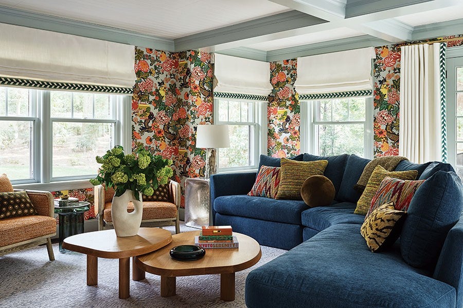 Living room with a large blue sectional sofa adorned with various patterned throw pillows, two wooden coffee tables with a vase of green flowers and books, floral wallpaper, three windows with white Roman shades, and two orange armchairs with patterned cushions. A silver floor lamp stands between the sofa and armchairs.