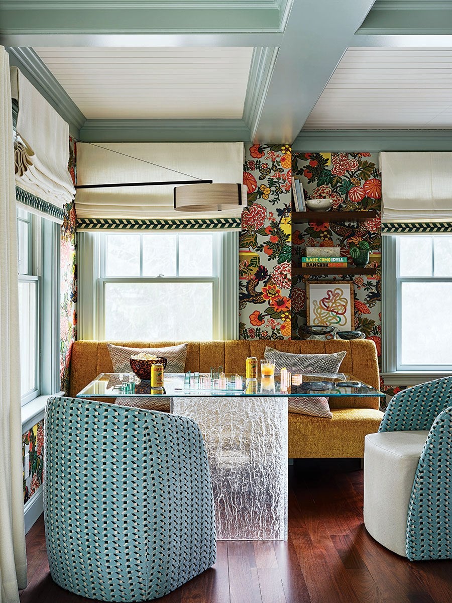 A cozy dining nook features a mustard yellow upholstered bench with patterned throw pillows against a vibrant floral wallpaper. A glass-top table with a textured, clear base sits in front of the bench, accompanied by two rounded chairs upholstered in a blue and white geometric fabric. The space is framed by light blue window trim and white Roman shades with a green chevron border. A modern, horizontal pendant light hangs above the table, and wooden floating shelves with books and decorative items are mounted on the wallpapered wall. The floor is dark wood.
