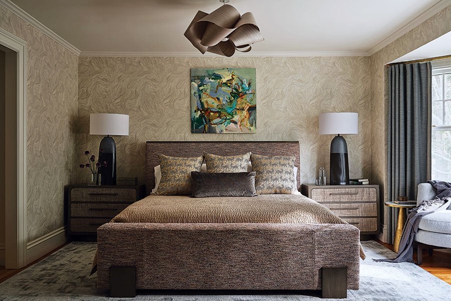 A modern bedroom featuring a large upholstered bed with a textured brown bedspread and five decorative pillows in neutral tones. On either side of the bed are matching dark wood nightstands with three drawers, each topped with a tall black lamp with a white shade. Above the bed hangs an abstract painting with green, blue, and orange hues. The walls have a subtle textured beige wallpaper, and a sculptural wooden ceiling light fixture is centered above the bed. To the right, a window with gray curtains lets in natural light, next to a light gray upholstered chair with a dark throw draped over it. A small round wooden side table sits beside the chair. The floor is covered with a light gray area rug.