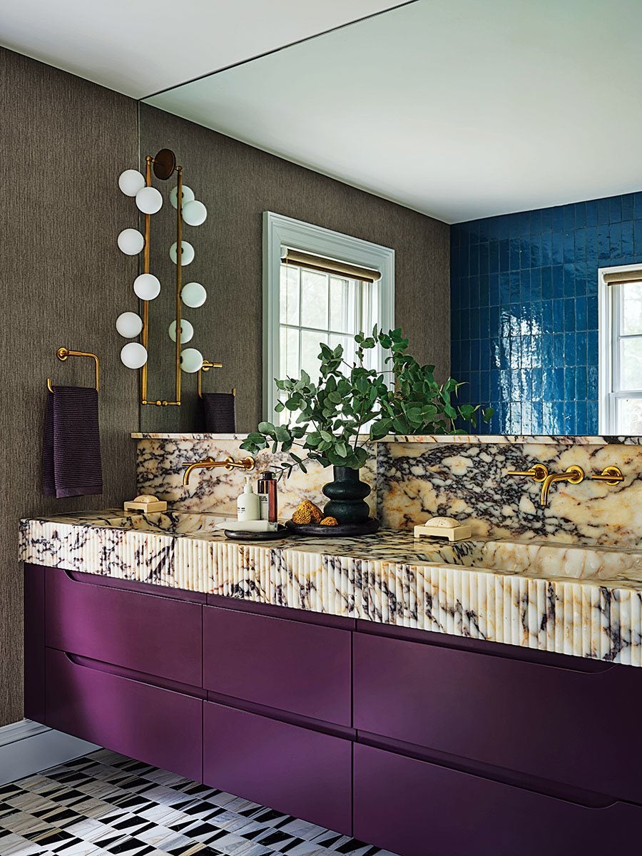 Bathroom vanity with a purple base and a marble countertop featuring intricate veining. The wall behind the vanity has a large mirror and two windows with white frames. The wall to the left is covered in textured brown wallpaper, while the wall to the right is tiled with glossy blue tiles. Gold fixtures include wall-mounted faucets and towel holders, with a dark towel hanging on the left. A black vase with green foliage sits on the countertop, alongside soap and toiletries. The floor has a geometric black, white, and gray tile pattern.