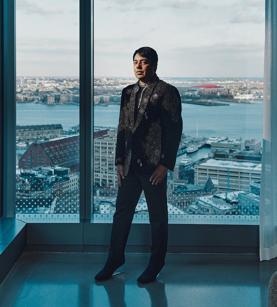 A man stands indoors in front of a large window with a cityscape and waterfront view. He is wearing a dark, patterned blazer, dark pants, and black shoes. The lighting highlights his face and upper body, and the background shows buildings, water, and a partly cloudy sky.