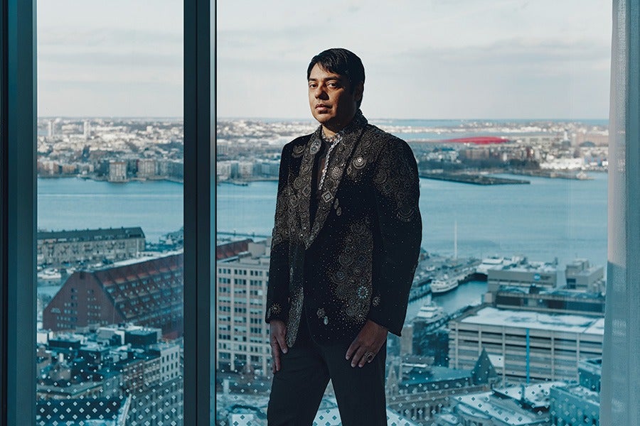 A man wearing a dark, patterned suit stands indoors in front of large windows overlooking a cityscape with water, buildings, and boats. The sky is partly cloudy, and the overall lighting is natural.