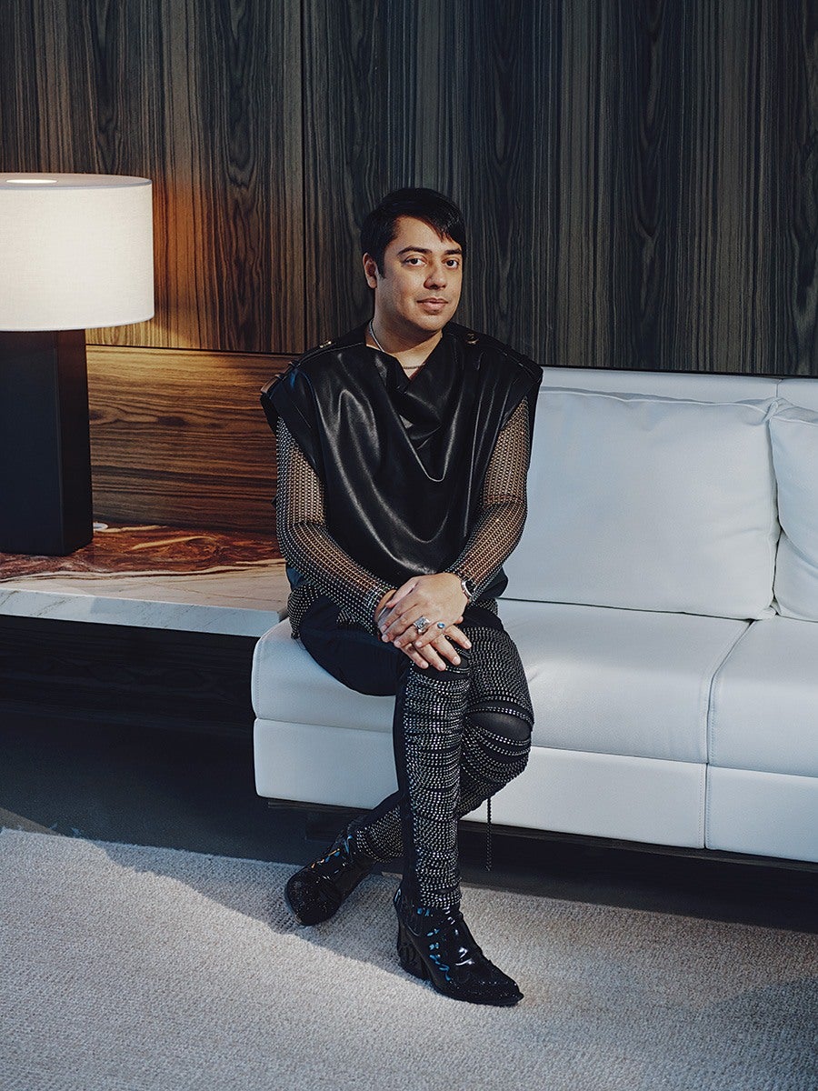A man is sitting on a white leather sofa with his legs crossed. He is wearing a black leather sleeveless top over a mesh long-sleeve shirt, paired with black pants adorned with silver studs or rhinestones. He has black shiny boots and is accessorized with rings and a watch. The background features dark wood paneling and a side table with a modern lamp. The floor is carpeted in a light color.
