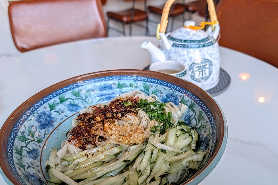 A bowl of noodles topped with chopped green onions, minced garlic, chili flakes, and shredded cucumber, served in a decorative blue and white floral bowl. In the background, there is a white and green patterned teapot with a bamboo handle and a matching teacup on a white marble table.