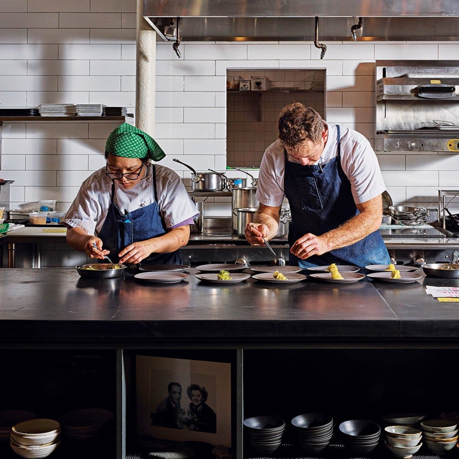 Two chefs wearing white shirts and blue aprons are carefully plating food in a professional kitchen. The chef on the left wears glasses and a green headscarf, while the chef on the right has short curly hair. They are working at a black countertop with several plates arranged in front of them. The background features white tiled walls, stainless steel kitchen equipment, and shelves with stacked plates and bowls.